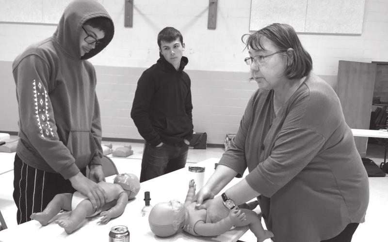Timber Lake EMT Kendra Enright (RIGHT) instructs high school students Aidan Rough Surface and Carter Keller in how to perform life-saving CPR on babies. The training is part of the health curriculum at Timber Lake School. Such classes contribute to the knowledge/skill base that can potentially prevent ambulance calls and exposes students to the possibility of becoming EMTs. Photo by Kathy Nelson