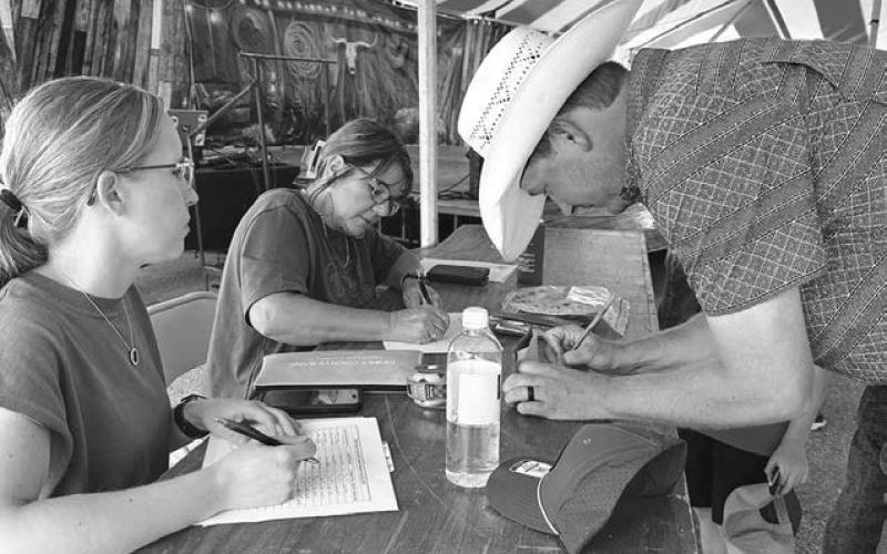Volunteers Kyrie Lemburg (left) and Rita Long recorded bids and collected money when cookies, pies and a Centennial belt buckle were auctioned off after the free supper provided by the Timber Lake Rodeo Association and Western Dakota Bank. Jace Parker (right) was one of the bidders who wrote a check. Auction proceeds went to Dewey County 4-H and the Rodeo Association.