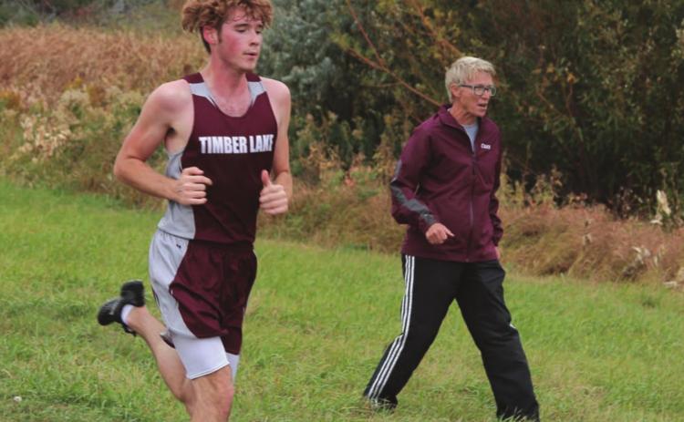 Timber Lake senior Ethan Farlee runs the homestretch of the Panthers’ home meet while coach Donna Aberle encourages him. Photo by Jon Flatland